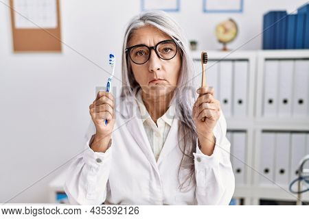 Middle age grey-haired woman working at dentist clinic holding electric and recycled teethbrush depressed and worry for distress, crying angry and afraid. sad expression. 
