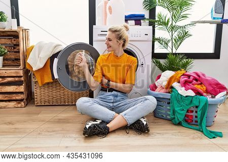 Young blonde woman doing laundry sitting by washing machine looking proud, smiling doing thumbs up gesture to the side 