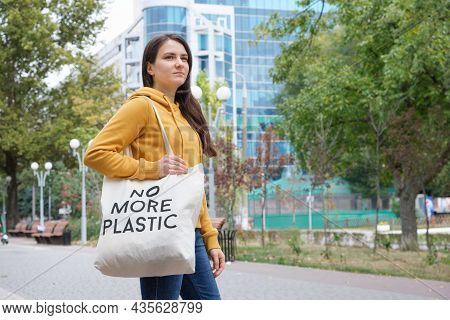 A Brunette Woman In An Orange Hoodie Walks With A Fabric Bag That Says No More Plastic In The City