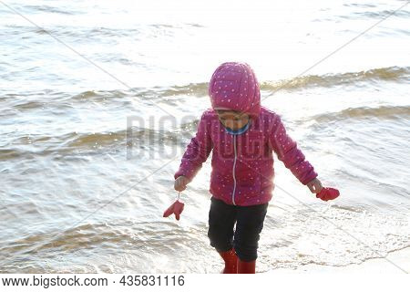 Child Girl Playing By The Sea In Autumn. A Girl In A Red Jacket In A Beautiful Autumn On The Seashor