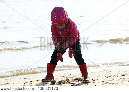 Child Girl Playing By The Sea In Autumn. A Girl In A Red Jacket In A Beautiful Autumn On The Seashor