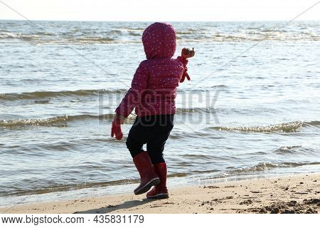 Child Girl Playing By The Sea In Autumn. A Girl In A Red Jacket In A Beautiful Autumn On The Seashor
