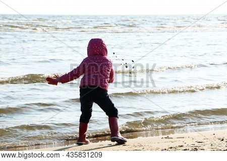 Child Girl Playing By The Sea In Autumn. A Girl In A Red Jacket In A Beautiful Autumn On The Seashor