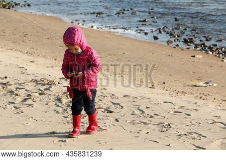 Child Girl Playing By The Sea In Autumn. A Girl In A Red Jacket In A Beautiful Autumn On The Seashor