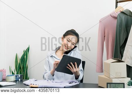 Asian Entrepreneur Talking On The Phone With A Customer While Checking Inventory On A Tablet.