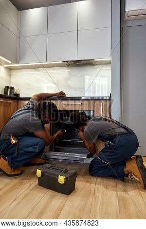 Two Experienced Repairpersons, Man And Woman, Fixing Kitchen Oven, Kneeing On Wooden Floor. Young Af