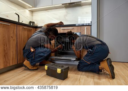 Back View Of Woman And Man In Overalls, Kneeling In Front Of Opened Kitchen Oven. African American R