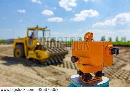 Surveyor Instrument For Measuring Level On Construction Site, Roller With Spikes Is Compacting Soil 