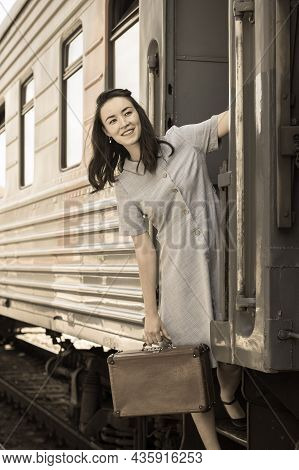 A Cheerful Woman With A Vintage Suitcase In Her Hand Looks Out Of The Train. Young Happy Woman Pulli