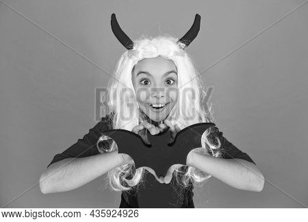 Funny Kid In Witch Costume Hat For Halloween With Devil Hors And Wig Holding Bat On Orange Backgroun