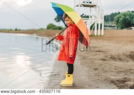 Boy In A Red Raincoat And Yellow Rubber Boots Stands At River Bank And Holding Rainbow Umbrella. Sch