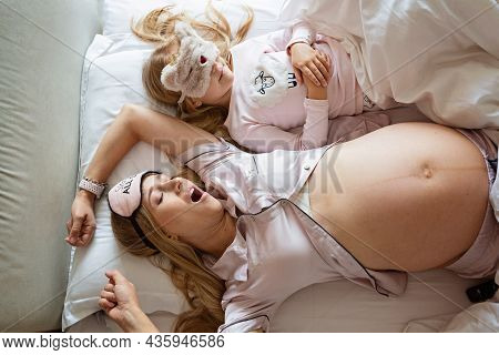 High View Of Pregnant Caucasian Mother With Big Belly And Daughter Sleeping In Blindfolds On The Bed