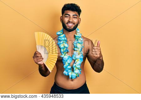 Arab man with beard wearing hawaiian lei holding handfan smiling happy and positive, thumb up doing excellent and approval sign 