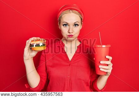 Young blonde woman eating a tasty classic burger with fries and soda puffing cheeks with funny face. mouth inflated with air, catching air. 