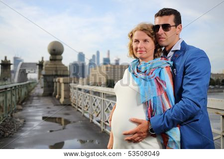 Pregnant woman in white and stylish man with hands on her belly look away on street.