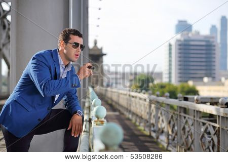 Handsome man in blue jacket and sunglasses with cigar stands on bridge and looks away.