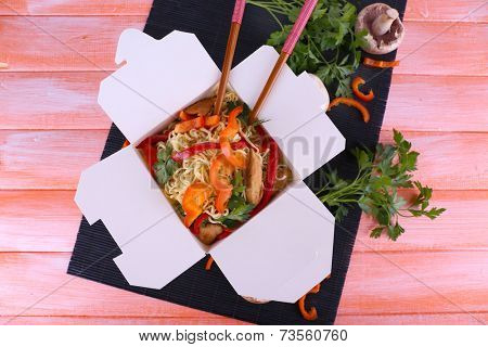 Chinese noodles and sticks in takeaway box on black mat on pink background
