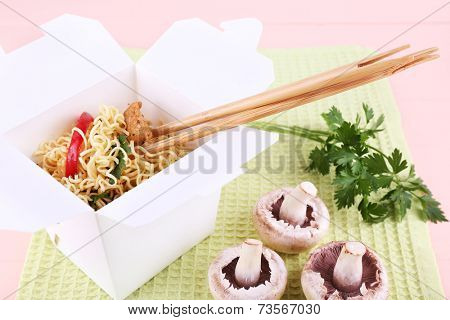 Chinese noodles and sticks in takeaway box on green napkin on pink background