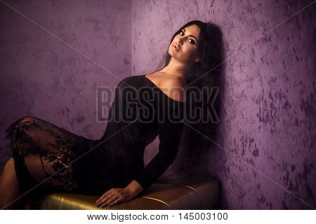 Portrait of sexy beautiful black-haired woman in high heels posing on metallic hassock in room with purple walls.