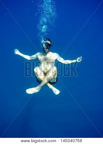 Portrait of a young causasian man meditating in the lotus position underwater. Submerged under water free diving crossed legs wearing a mask and blowing bubbles.