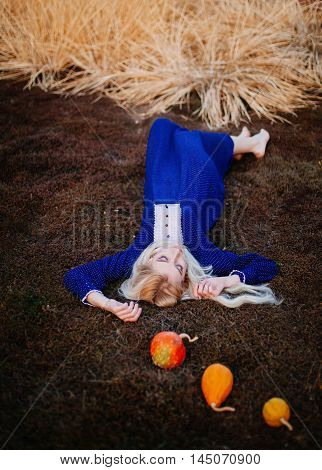 A woman dressed in a blue vintage dress lies in the autumn park. Small bright pumpkins lie next to her head.