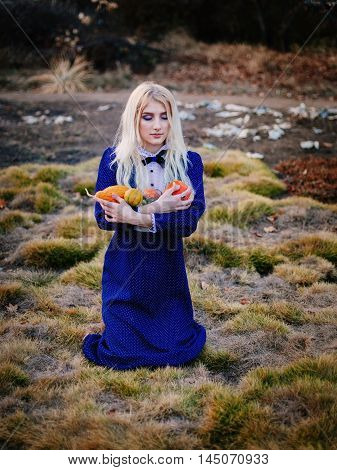A woman dressed in a blue vintage dress sits in the autumn park. She holds a small bright pumpkins.