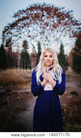 A woman dressed in a blue vintage dress stays in the autumn park. It`s cold there.