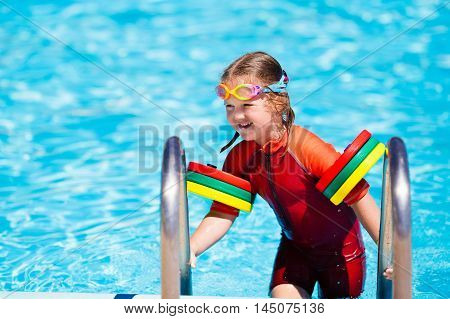 Happy laughing little girl playing in outdoor swimming pool on a hot summer day in sun protection rash guard. Kid in goggles and UV safe bathing suit learning to swim with colorful floaties.