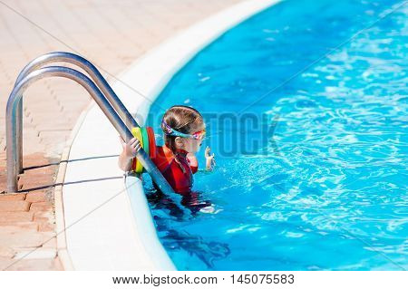 Happy laughing little girl playing in outdoor swimming pool on a hot summer day in sun protection rash guard. Kid in goggles and UV safe bathing suit learning to swim with colorful floaties.