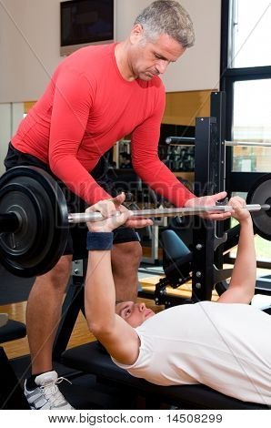 Young man doing weigths on a bench press with the help of the mature instructor at gym