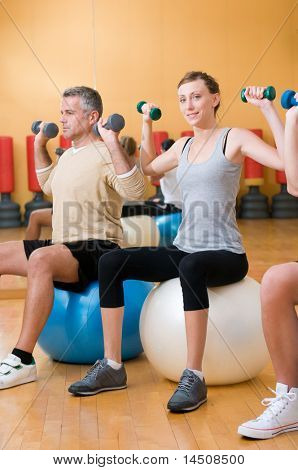 Young lady smiling and looking at camera while lifting weigths on a fitness ball at gym