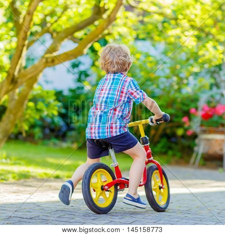 funny blond kid boy in colorful clothes driving bicycle in domestic garden. Toddler child dreaming and having fun on warm summer day. Active games for children outdoors.
