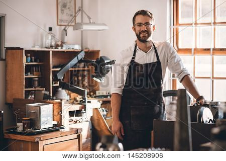 Portrait of a smiling young jeweler standing in a workshop full of tools and jewelry making equipment