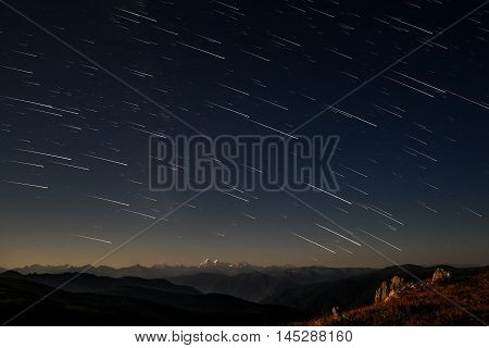 Beautiful night landscape with traces of stars in the form of tracks in the night sky against the background of the contours of mountains and snowy peaks shot with a long exposure