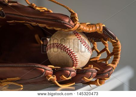 Close-up of a used baseball ball inside brown leather baseball glove on plain grey background.