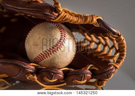 Close-up of a used baseball ball inside brown leather baseball glove on plain grey background.