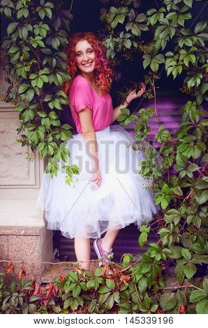 Happy smiling beautiful young woman with curly red hair with colorful hair pieces wearing bouffant skirt and magenta blouse standing among grape leaves