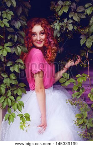 Happy smiling beautiful young woman with curly red hair with colorful hair pieces wearing bouffant skirt and magenta blouse standing among grape leaves
