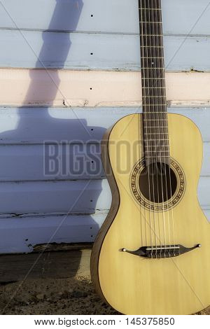 Parlour-sized classical acoustic guitar resting against a beach hut painted in pastel pink and blue. Symbolic of vacation and beach relaxation.