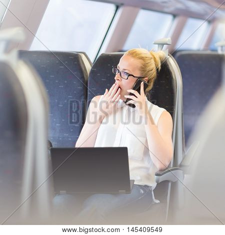 Businesswoman traveling by train, yawning while talking on cellphone and working on laptop. Long and tiresome business travel concept.