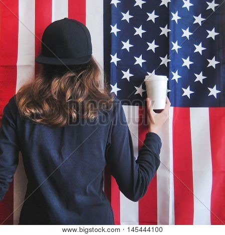 Vintage lifestyle portrait of young hipster girl holding white paper cup of coffee against american flag background dressed in casual clothes black pullover and snapback cap. USA love for coffee concept