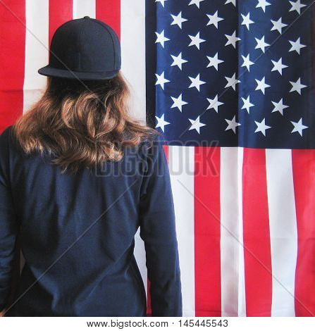 Bright colorful lifestyle portrait of young hipster girl turned back on american flag background dressed in casual clothes black pullover and black snapback cap with hair down