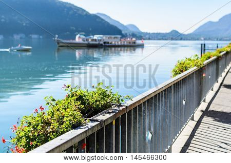 Vacation summer sunny lake view. Beautiful flowers over the background of beautiful serene blue Lugano lake clear blue sky and a ship surrounded by hills in Morcote Switzerland selective focus