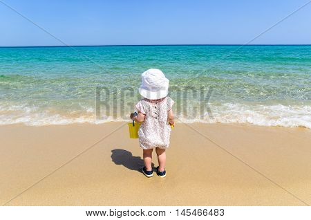 Adorable little girl in beautiful swimsuit and white hat holding kid bucket and having fun at exotic white sandy beach during vacation over the background of turquoise sea and blue sky copy space