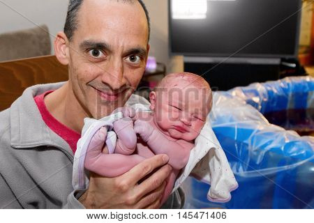A proud father holds his brand new son up for a picture. The birthing pool that the baby was just born in is in the background.