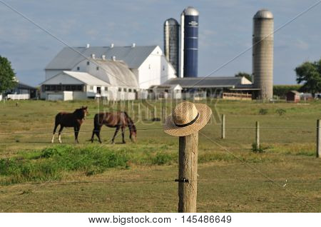 Amish straw hat laying over fence post with Amish farm and horses in background
