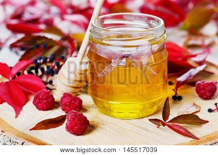 Sweet honey in pot with bow on a wooden background decorated autumn leaves and raspberry, focus on the front of the pot.