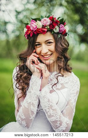 Wedding. Young beautiful bride in white dress and wreath standing at green lawn between trees. Summer full length atmospheric portrait portrait.