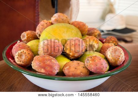 Prickly pear cactus fruits and mango fruits in the plate on the table on the background a chair with a straw hat on it. Mango and prickly pear cactus fruits in a plate. Horizontal. Close.