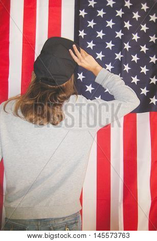 Bright colorful lifestyle portrait of young hipster girl taking off cap turned back on american flag background dressed in casual clothes gray sweater and black snapback cap with hair down